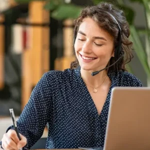 lächelnde Frau vor einem Laptop, trägt Kopfhörer und macht sich Notizen