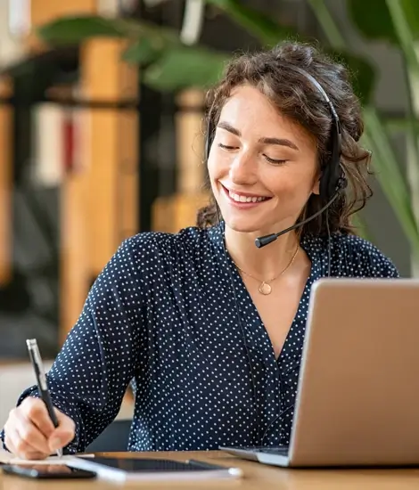 lächelnde Frau vor einem Laptop, trägt Kopfhörer und macht sich Notizen